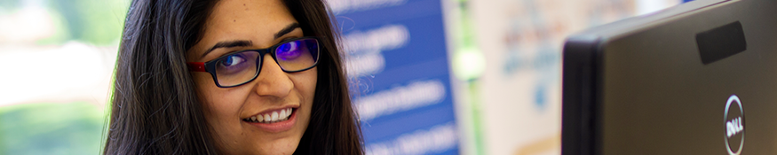 A female student with glasses, smiling, sat in front of a PC monitor